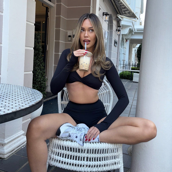 Woman in black outfit sitting on a white chair outdoors, holding a drink.